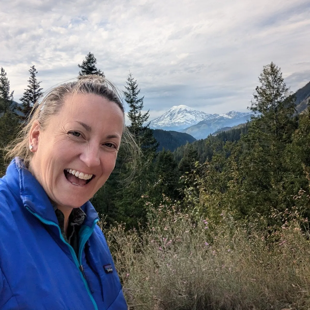 Elizabeth Sall laughing on a mountain trail, with evergreens and a snowy Cascade peak behind her